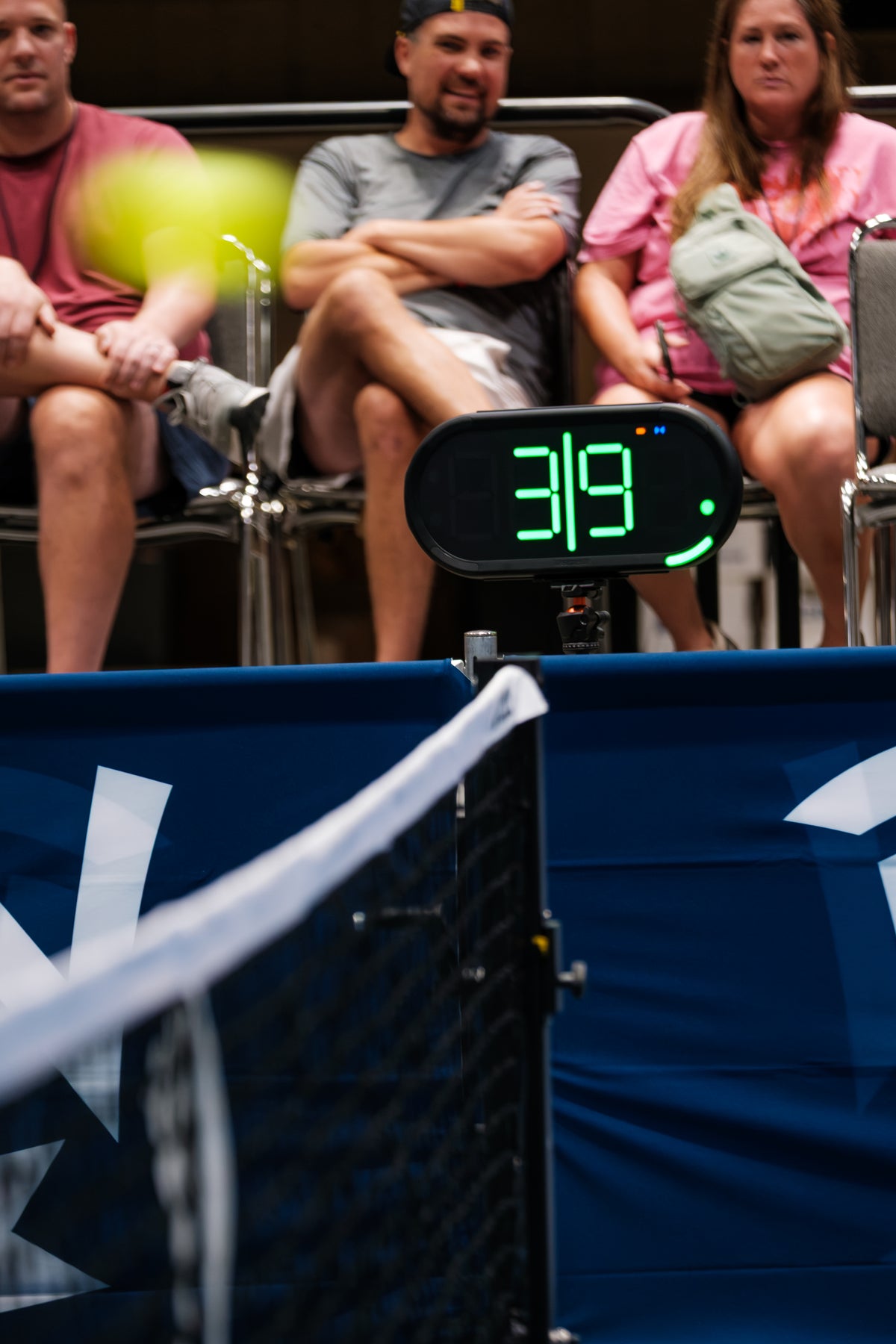 A Dropshot scoreboard showing 3|9 server 1 from the left side, with a blurred pickleball in the foreground and spectators seated behind a blue barrier at an event.