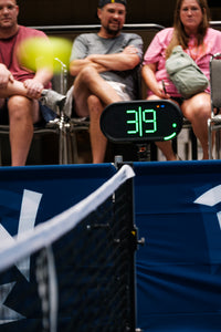 A Dropshot scoreboard showing 3|9 server 1 from the left side, with a blurred pickleball in the foreground and spectators seated behind a blue barrier at an event.