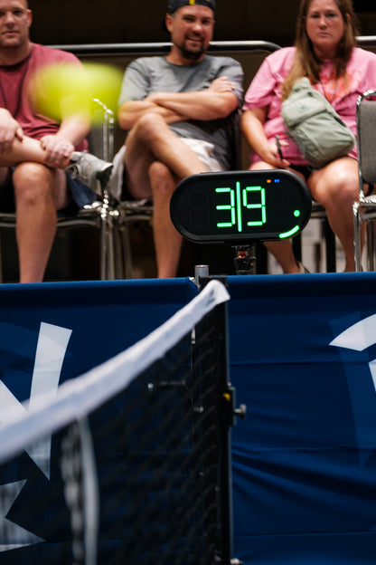 A Dropshot scoreboard showing 3|9 server 1 from the left side, with a blurred pickleball in the foreground and spectators seated behind a blue barrier at an event.