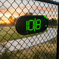 A Dropshot scoreboard with bright green LED numbers showing '10|8' server 1 from the right service area on a chain-link fence, with a sunset and outdoor field visible in the background.