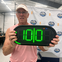 A man in a pink shirt and beige cap holds up a Dropshot scoreboard displaying the number 10|0 server 1 from the right side in bright green LED digits. 