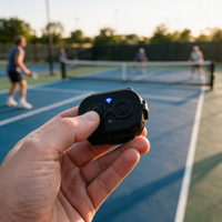 A hand holds a small black wireless remote control in the foreground, with a blurred pickleball court and players in the background during golden hour.